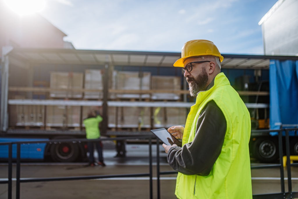 Warehouse manager overseeing unloading of truck, holding tablet, looking at cargo details, checking delivered items, goods against order.