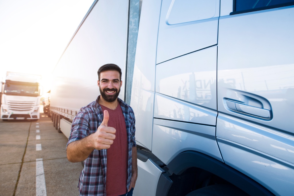 Retrato de joven barbudo con el pulgar hacia arriba de pie junto a su camión. Conductor de camión profesional y positivo de pie junto a un vehículo semirremolque. Servicios de transporte.