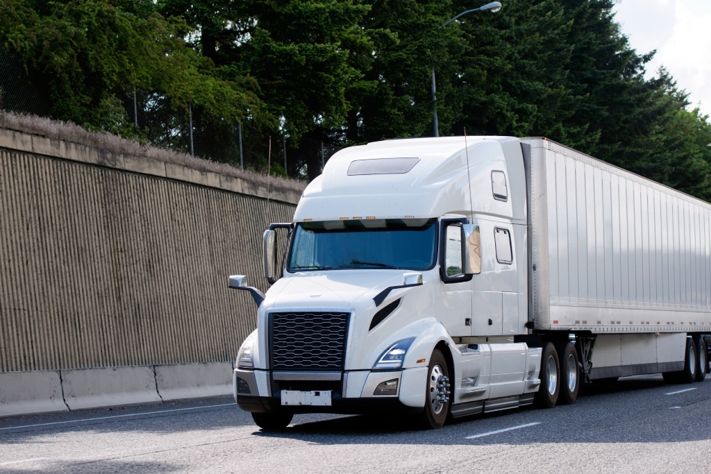 Elegant powerful white modern bonnet big rig long haul semi truck transporting commercial cargo in dry van semi trailer on the road with concrete side wall and trees on the hill